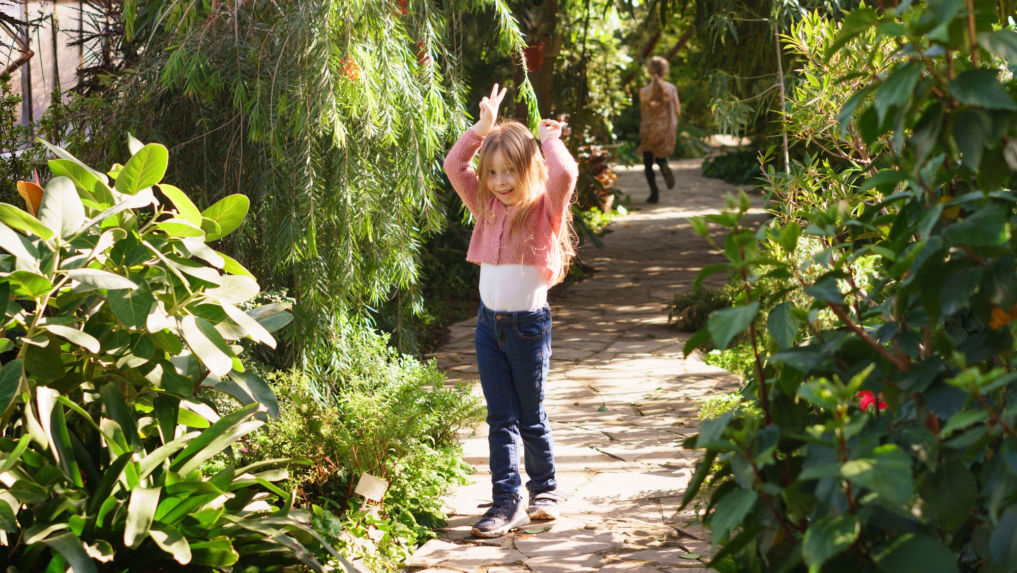 A young girl stands on a sunlit garden path surrounded by lush green plants, wearing a pink cardigan, white top, and blue jeans, with her hands raised in the air. Another person is visible in the background of the garden.